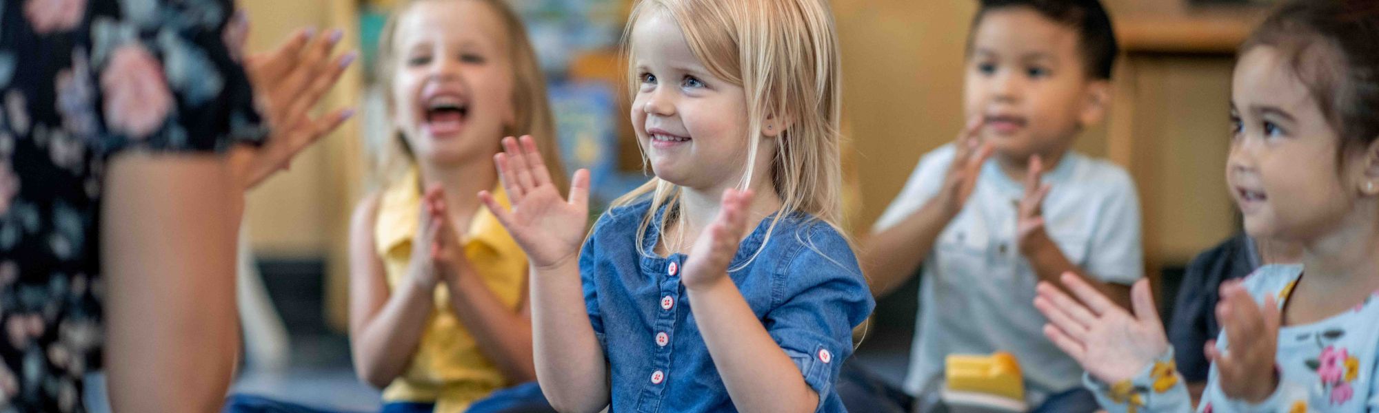 A group of young children sit in a classroom circle, smiling and clapping their hands while engaging with an adult leading an activity. The children appear happy and attentive, suggesting a positive, interactive learning environment focused on connection and participation.
