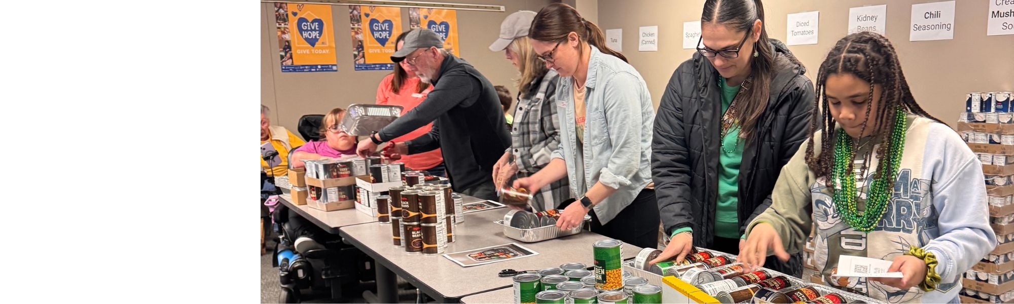 A group of volunteers stands around tables assembling meal kits with canned goods and packaged food items. People of different ages work together, organizing and placing items into trays, with stacks of food and labeled ingredients visible in the background.